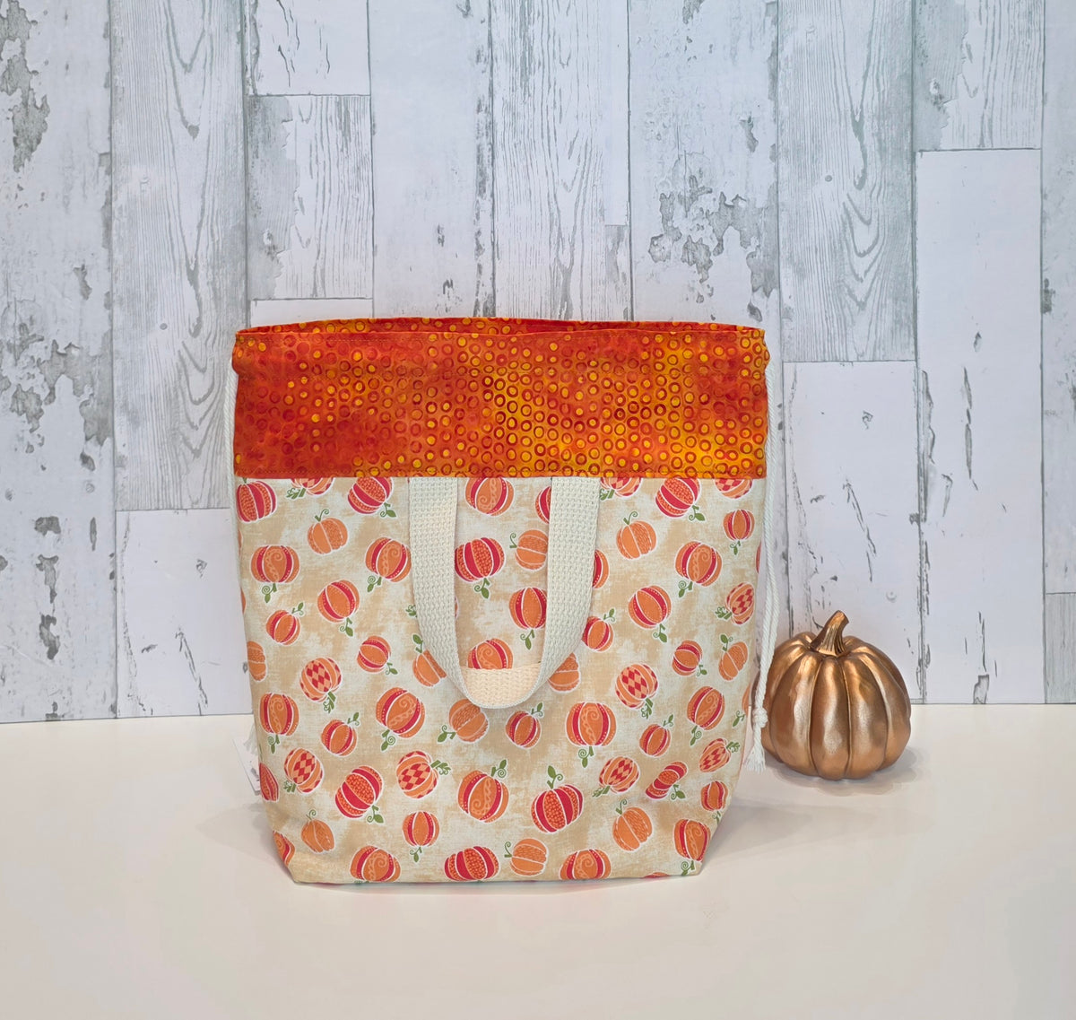 A fabric drawstring project bag with a pumpkin pattern on a cream background, featuring a bright orange and yellow sequined top border and white canvas handles, displayed next to a small metallic gold pumpkin on a white surface against a rustic gray wood plank background. This is the back view of the bag and shows the handle.