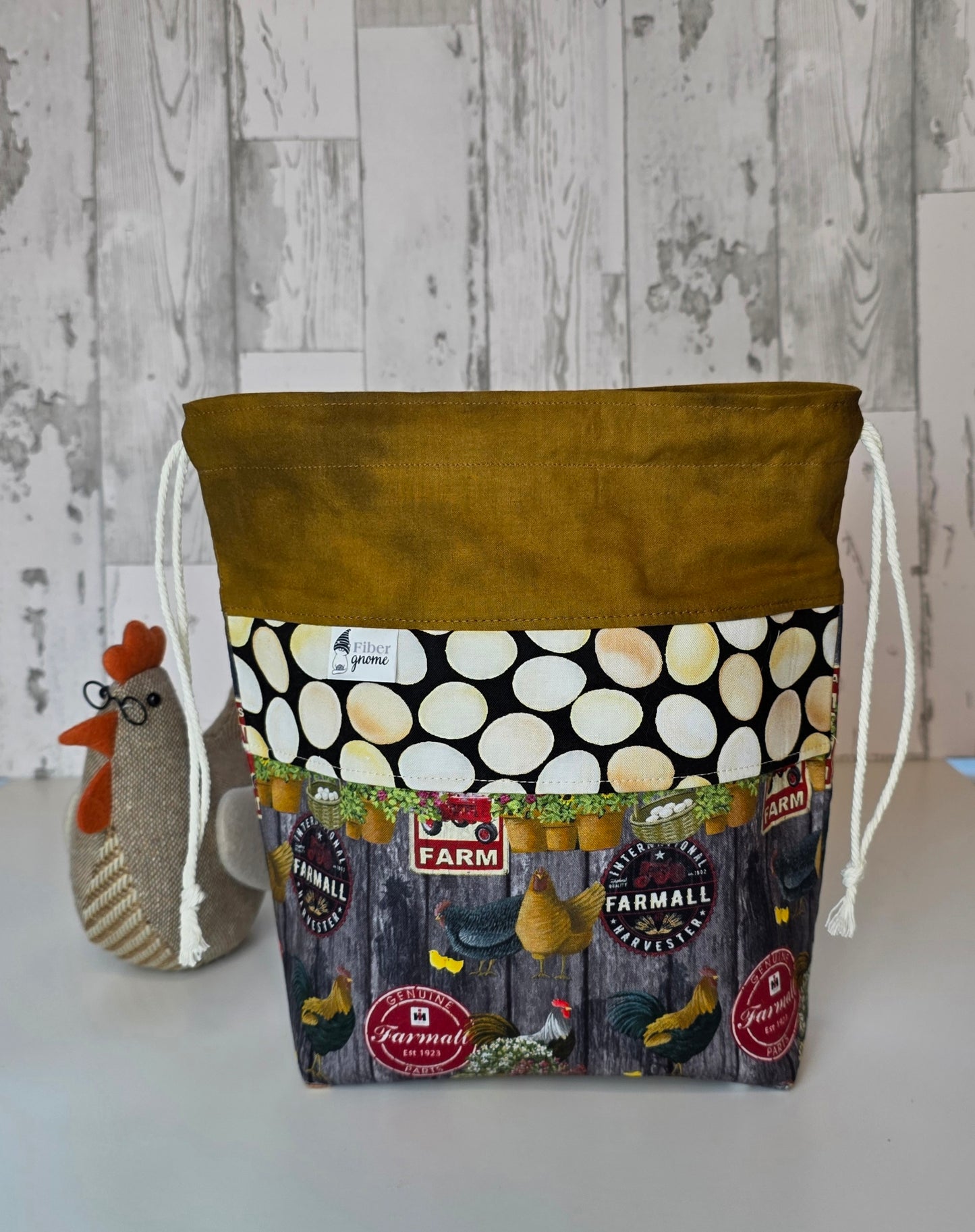 Fabric drawstring bag with golden brown contrast fabric, panel with egg design, and main fabric with chickens and labels. Stuffed chicken for scale and white wooden background.