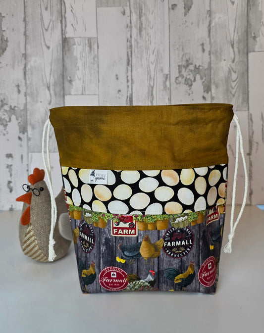 Fabric drawstring bag with golden brown contrast fabric, panel with egg design, and main fabric with chickens and labels. Stuffed chicken for scale and white wooden background.