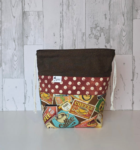 Drawstring bag with brown contrast fabric, panel in rusty red with cream dots, main fabric has vintage coffee labels. White wood background.