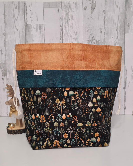 Drawstring bag in cotton fabric, top is in a golden wheat color, with a rustic teal panel and tree motif on the bottom portion. Wooden accent piece for scale and a faux wood background.