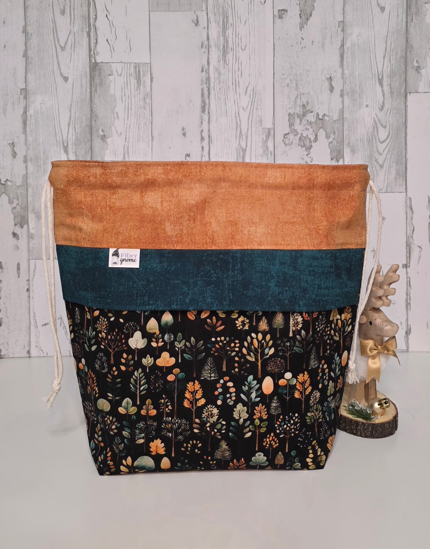 Drawstring bag with contrast fabric in golden wheat, accent panel in teal and main fabric is on a dark background with trees. Wood accent piece for scale and white wood background.