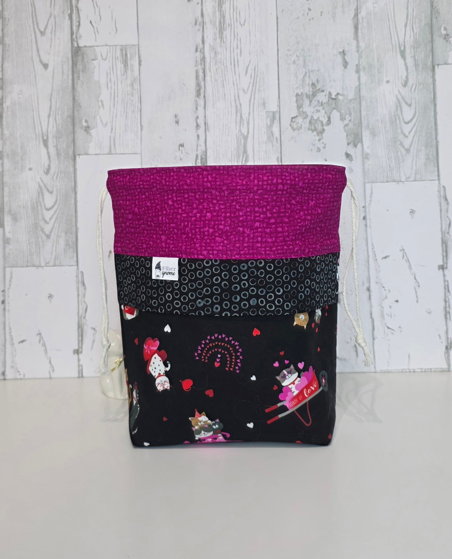 Fabric drawstring bag, contrast fabric in bright pin, accent panel in black with grey circles and main fabric with kittens and hearts. White wooden background.