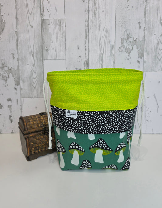 Fabric drawstring with bright green contrast fabric, black and white panel and main fabric with mushrooms in a dark green background. Wood box for scale and white wooden background.