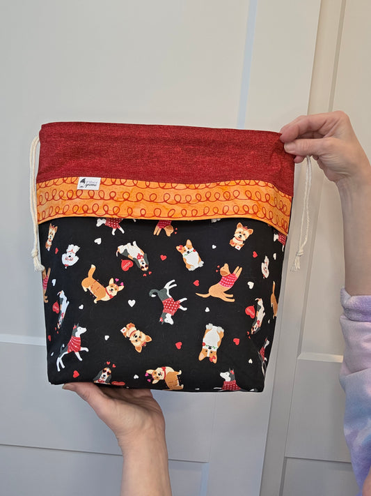 Drawstring bag with contrast fabric in red, an orange panel with red squiggles and the main fabric is a black background with dogs and hearts. White wood background.
