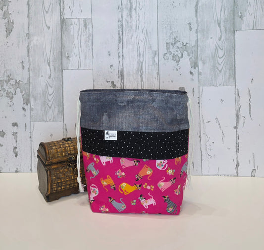 A fabric drawstring project bag, featuring a dark gray top band, a black polka-dot middle band, and a bright pink bottom panel printed with colorful cats in witchy like hats. It is displayed next to a small wooden chest against a rustic white wood plank background.
