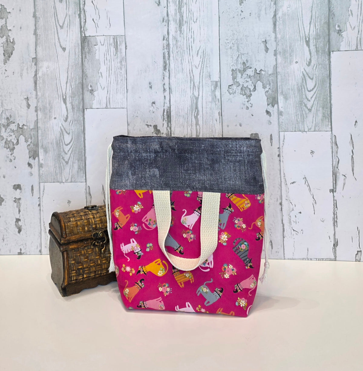 A fabric drawstring project bag, featuring a dark gray top band, and a bright pink bottom panel printed with colorful cats in witchy like hats. It is displayed next to a small wooden chest against a rustic white wood plank background. This is the back of the bag and shows the handle.