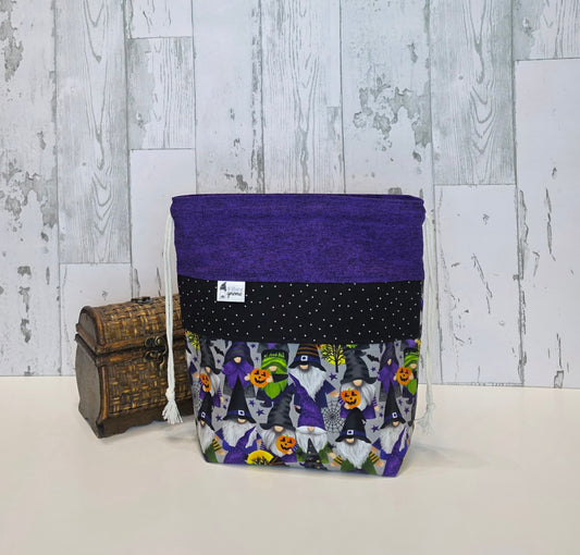 A fabric drawstring project bag with a purple top band, a black polka-dot middle band, and a Halloween-themed print lower section featuring witches, wizards, and pumpkins. It is displayed next to a small wooden chest against a rustic white wood plank background.