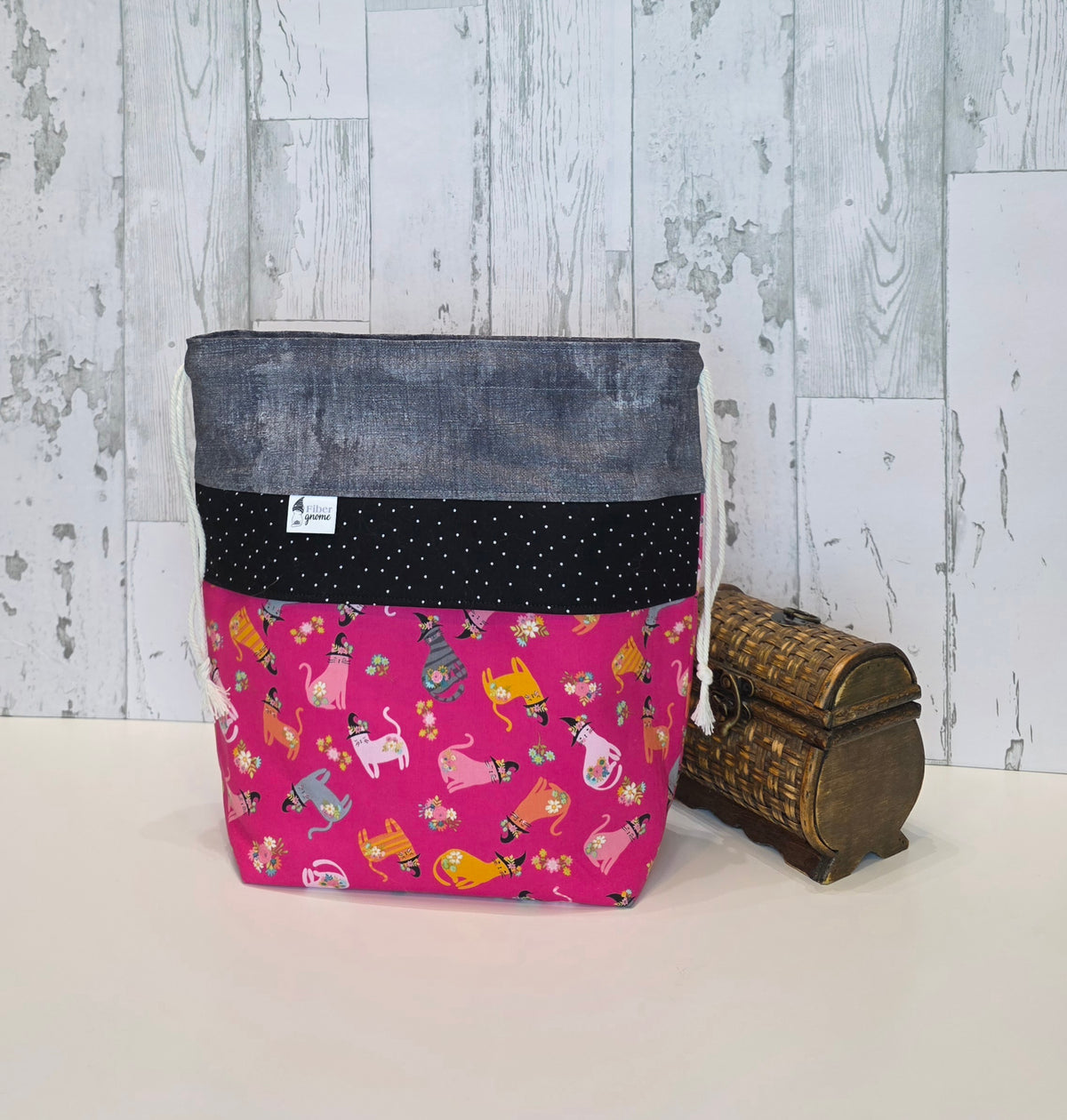 A fabric tote bag with a multi-patterned design, featuring a gray top section, a black polka-dot middle band, and a bright pink bottom panel with a whimsical cat print. It is displayed next to a small decorative wooden chest against a rustic white wood-paneled wall.