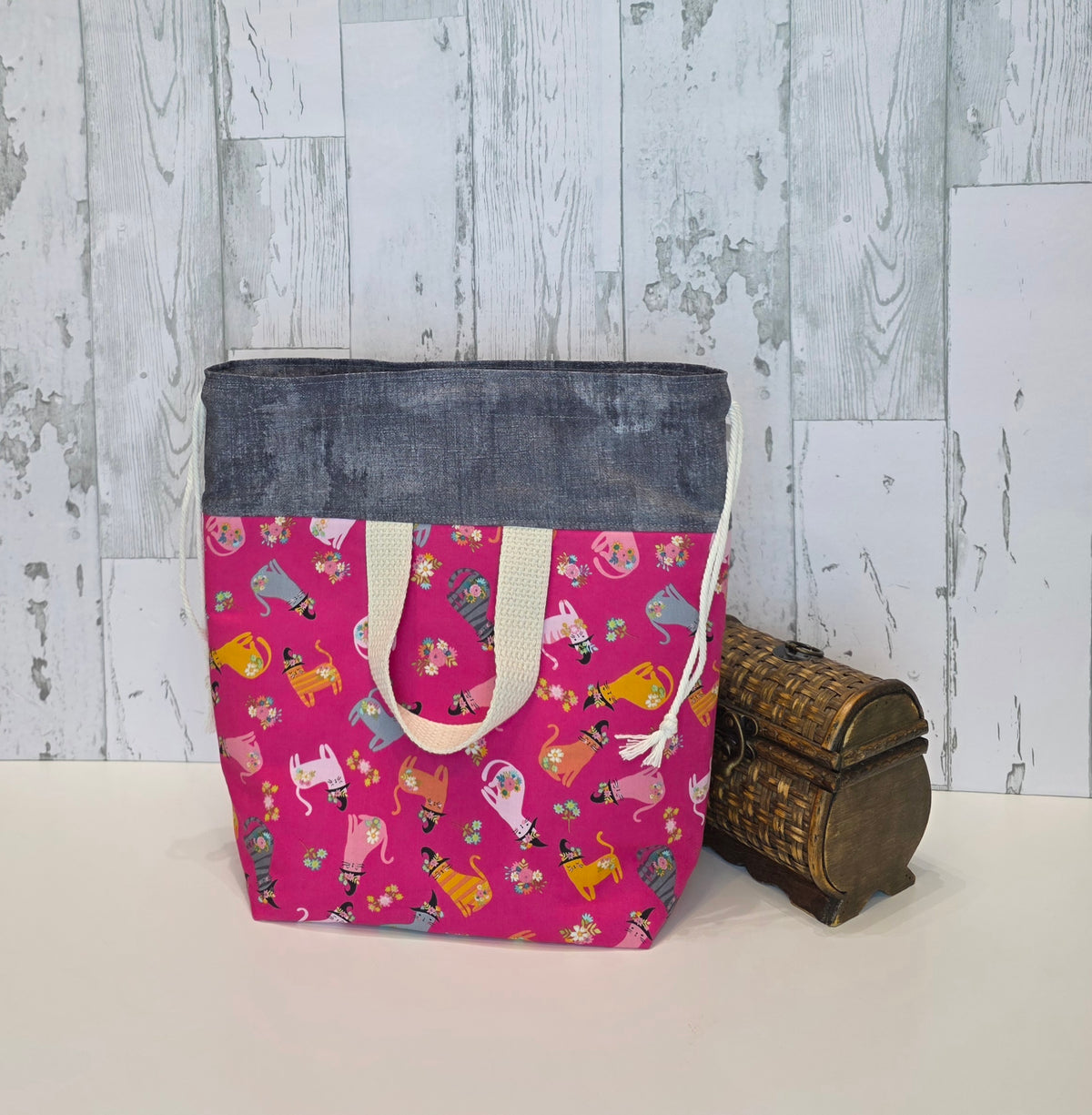 A tote bag with a bright pink patterned body, a dark gray denim-like top panel, and cream fabric handles, standing on a white surface against a light gray wood-plank backdrop. A small brown woven chest sits beside it as a prop. Back view of bag shows handle