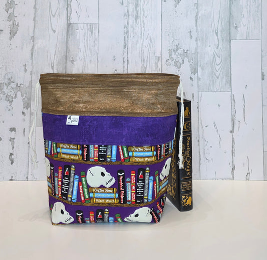 A fabric drawstring project bag with brown fabric top, a purple center panel and a printed bookshelf pattern featuring white skulls and colorful books, shown next to a vintage book on a white surface against a rustic white wood plank background.