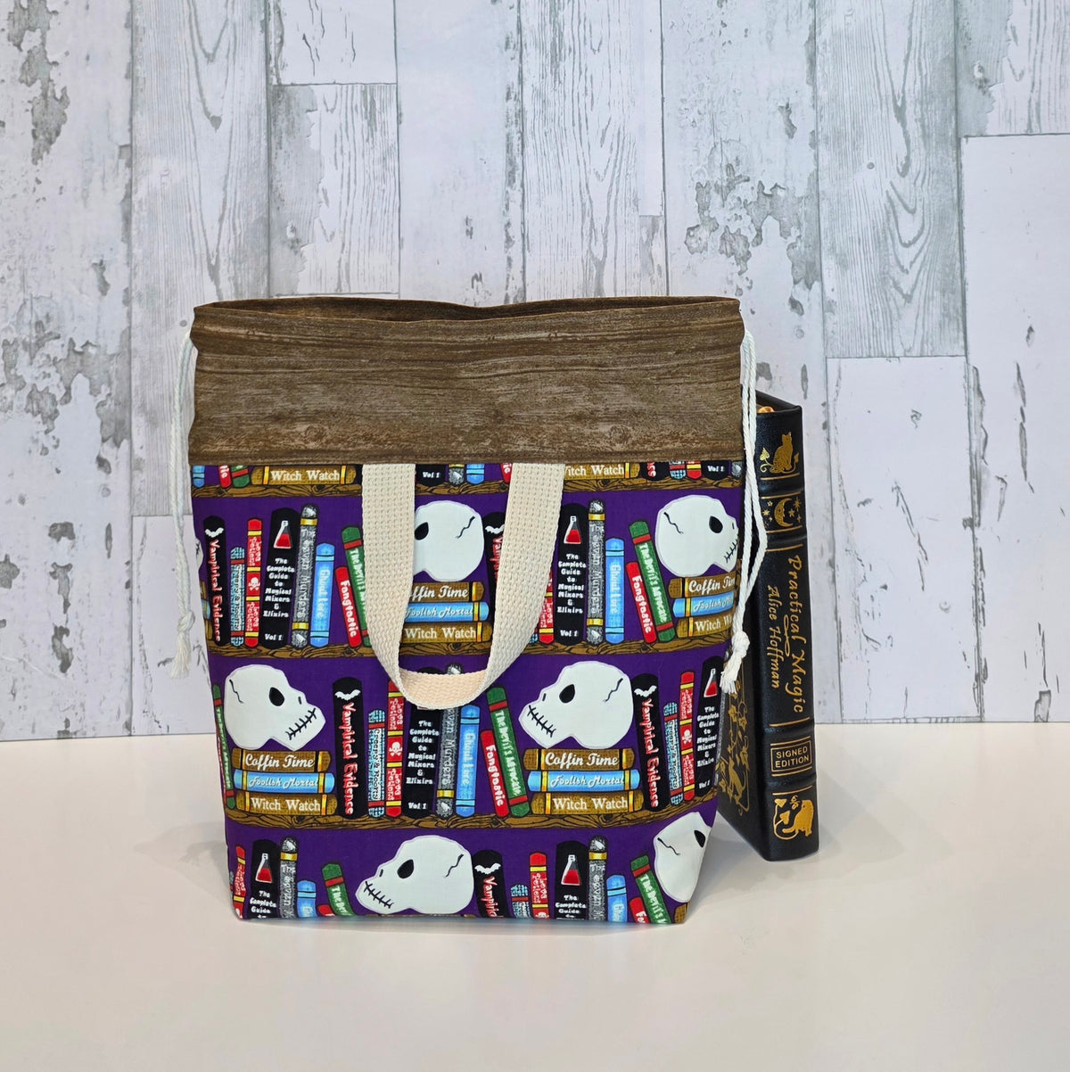 A fabric drawstring project bag with brown fabric top and a printed bookshelf pattern featuring white skulls and colorful books, shown next to a vintage book on a white surface against a rustic white wood plank background. This is the back view of the bag and shows the handle.