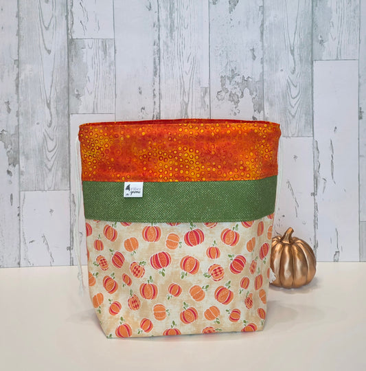 A fabric drawstring bag with a three-layered design: an orange patterned top, a green middle band, and a bottom section featuring an orange pumpkin print on a cream background. It is displayed on a white surface next to a small metallic gold pumpkin prop against a gray wood-plank background.