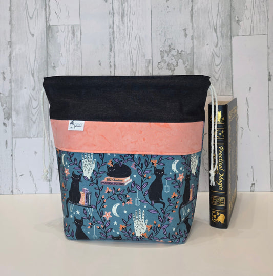 A fabric drawstring project bag with a black top section, a peach middle band, and a blue lower section printed with black cats, candles, and stars. It is shown standing on a white surface next to a hardcover book against a light gray wood-plank background.